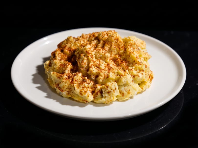 A white plate holds a serving of creamy potato salad, generously sprinkled with paprika.