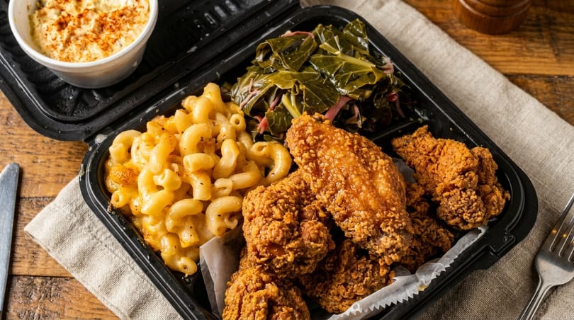 Fried chicken, macaroni and cheese, and collard greens in a black takeout container.