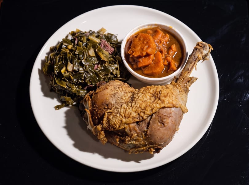 A plate of roasted duck leg, collard greens, and candied yams on a dark surface.