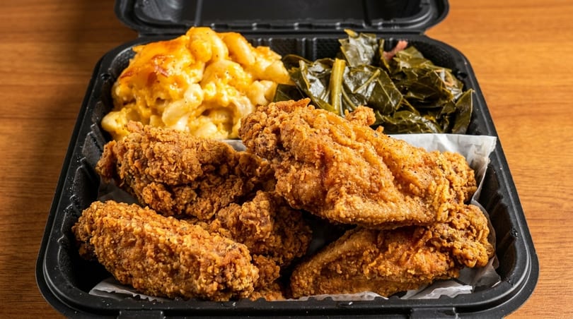 Fried chicken, macaroni and cheese, and collard greens in a black takeout container on a wooden table.