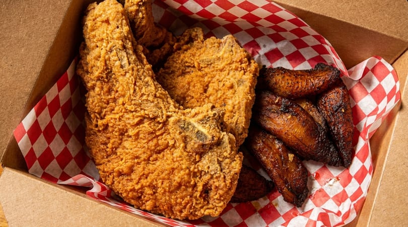 Crispy fried chicken and sweet plantains served in a cardboard box with red and white checkered paper.